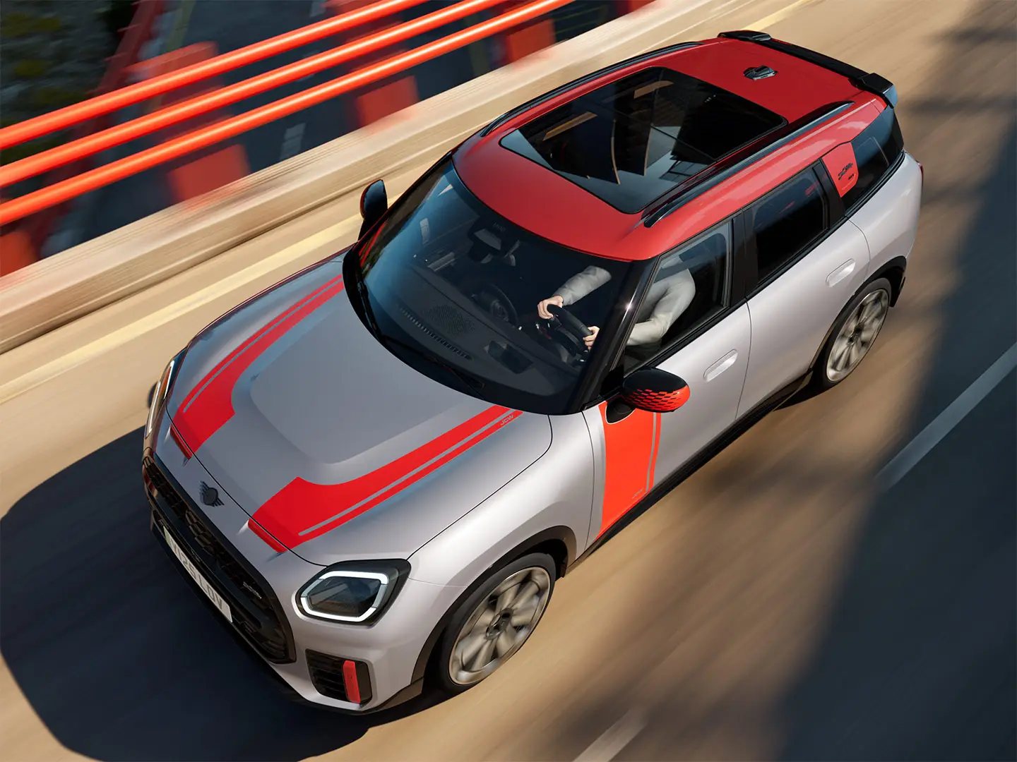 MINI Countryman John Cooper Works in Melting Silver with mirror cap, side and bonnet decals, roof, and winglet in Chili Red, driving along a city street – front-side view.