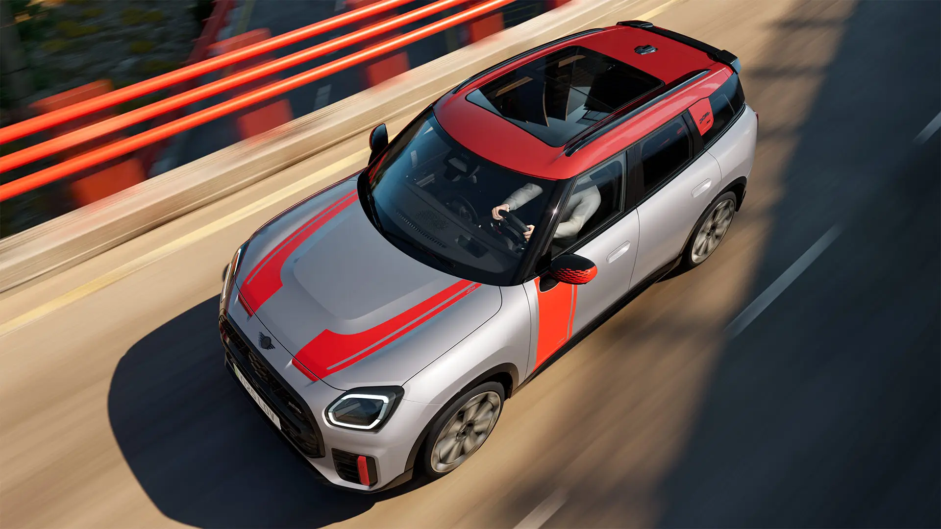 MINI Countryman John Cooper Works in Melting Silver with mirror cap, side and bonnet decals, roof, and winglet in Chili Red, driving along a city street – front-side view.