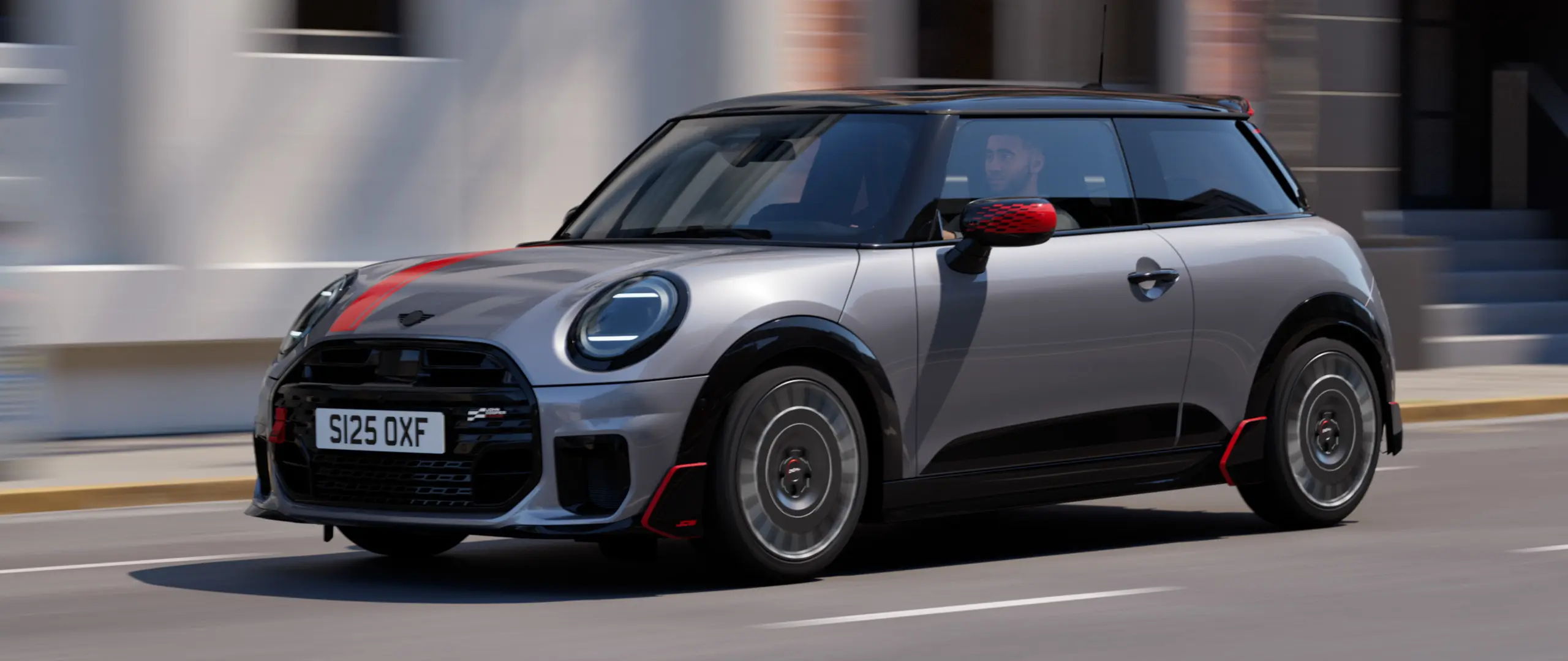 MINI Cooper 3-door in Melting Silver with John Cooper Works mirror cap and bonnet decal in Chili Red, roof spoiler and winglet in Black, driving along a city street – front-side view.