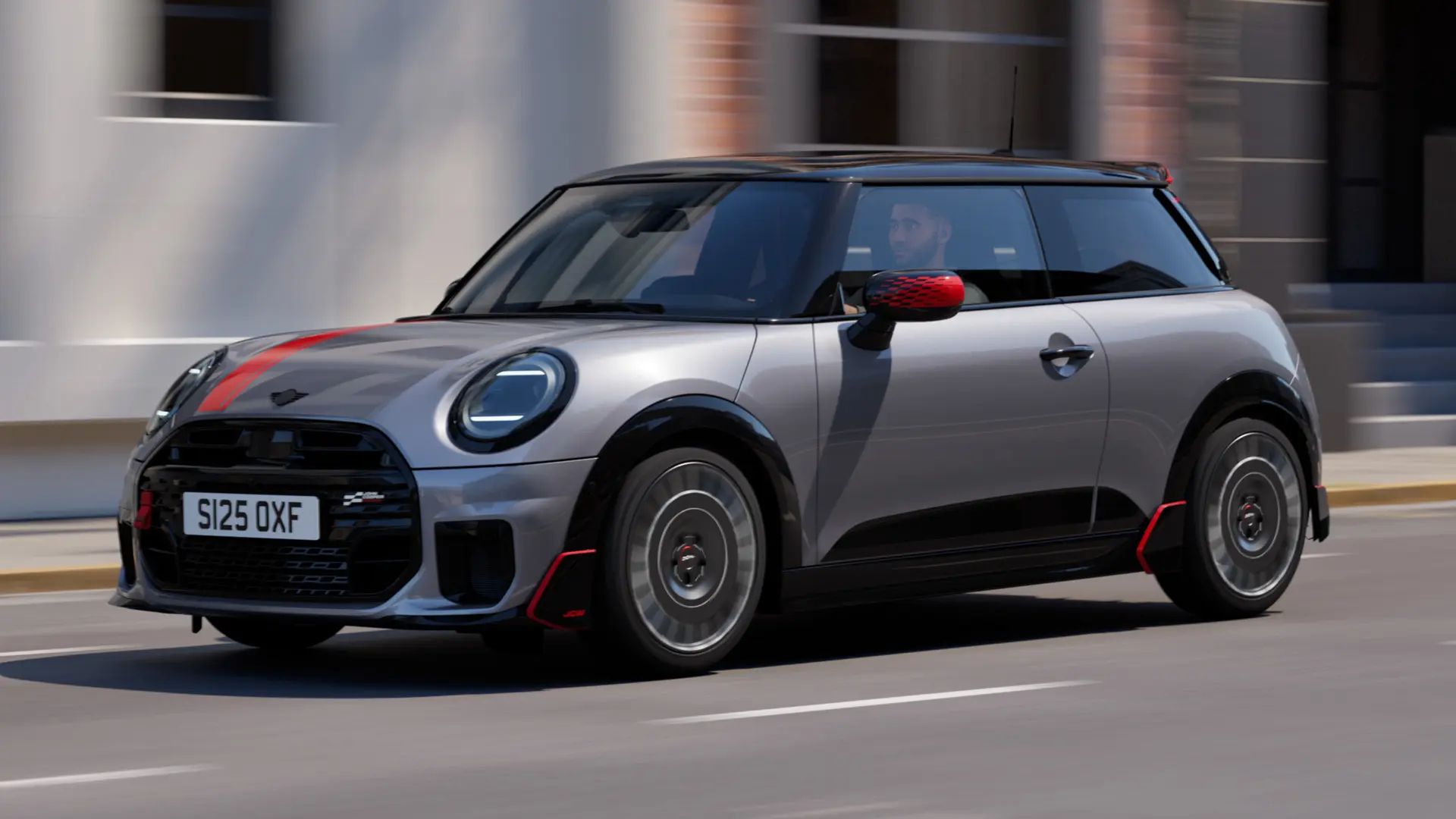 MINI Cooper 3-door in Melting Silver with John Cooper Works mirror cap and bonnet decal in Chili Red, roof spoiler and winglet in Black, driving along a city street – front-side view.