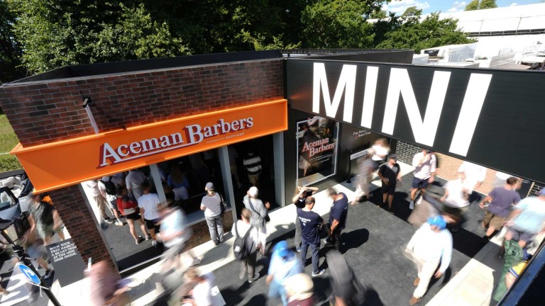 A bustling street scene outside a barbershop with a bright orange sign reading "Aceman Barbers." Crowds move energetically past, under a large "MINI" sign, suggesting a lively event.
