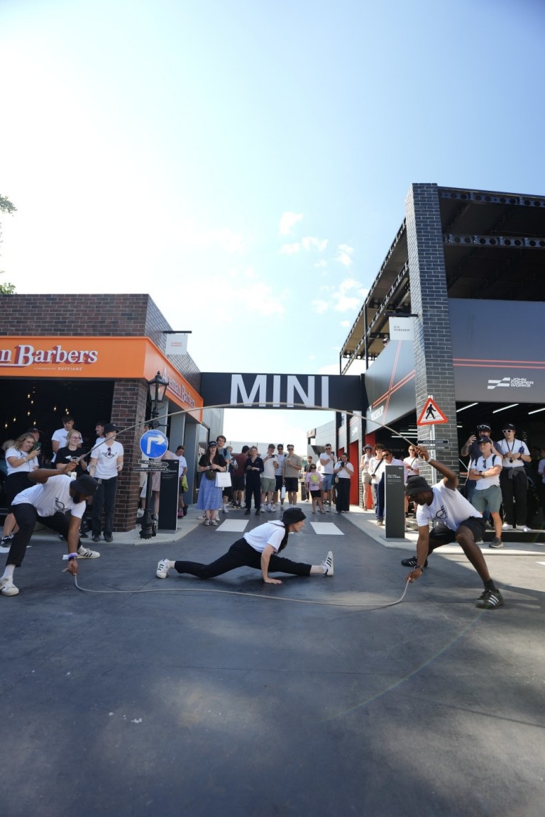  People watch performers doing a street dance routine in front of a MINI sign and brick storefronts, creating a lively and energetic atmosphere. 