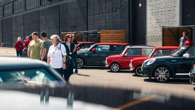 People gather outside a large black building, admiring a row of classic and modern Mini cars.