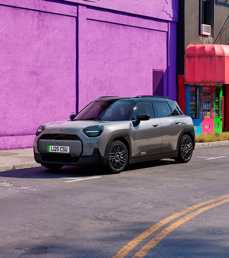 Gray MINI Aceman parked on a city street beside a bright purple and red building, featuring modern design and black roof.