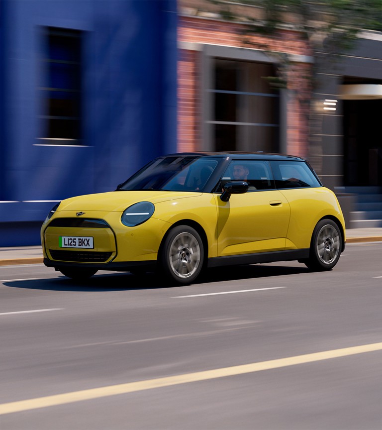 Yellow MINI Cooper electric hatchback driving on a city street with blue and brick buildings in the background, featuring black roof, circular LED headlights, and sleek modern design.