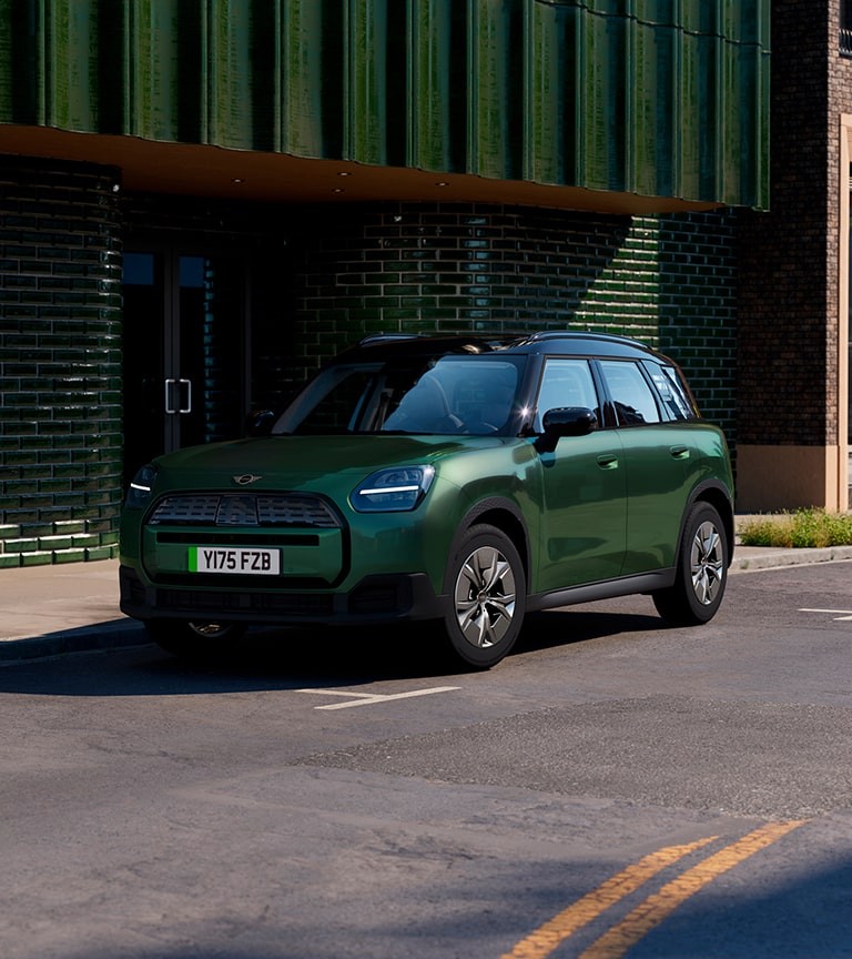 Green MINI Countryman electric SUV parked on a city street in front of a dark green tiled building, featuring black roof and modern headlights.