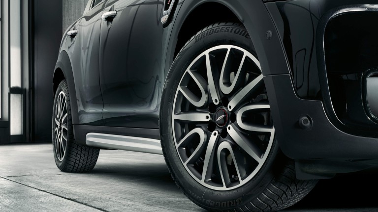 Close-up of a Mini Cooper Countryman wheel and tire, featuring stylish alloy rims and Bridgestone tires on a sleek black vehicle. The car is parked indoors on a concrete floor.