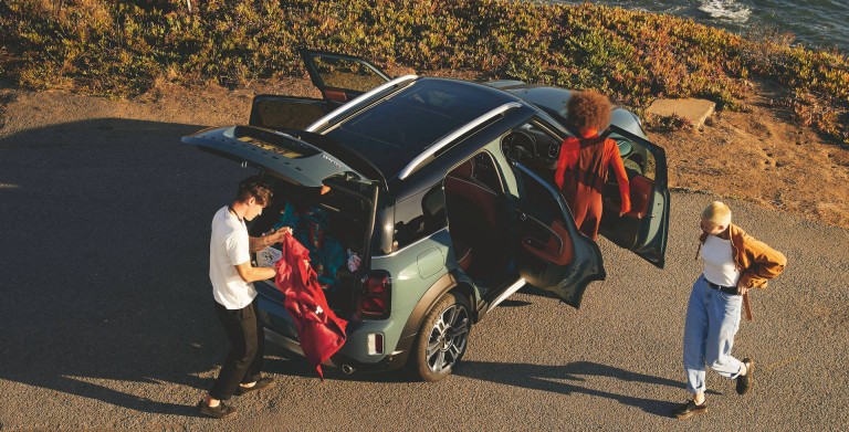 Three friends unpacking a green MINI Countryman parked near a coastal road at sunset. The car’s rear hatch and doors are open as one person retrieves clothes from the trunk.