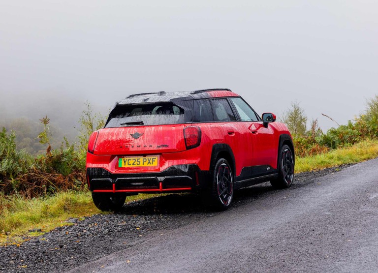 Rear side view of parked all-electric MINI Aceman in British Racing Green in car park with all doors open and coloured wall in the background